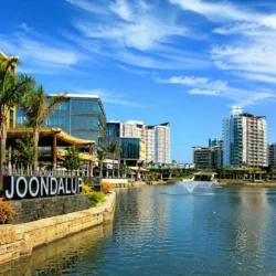 Modern waterfront cityscape of Joondalup, Perth, featuring a calm lake with fountains, contemporary buildings, palm trees, and a clear blue sky, with a visible “JOONDALUP” sign in the foreground.