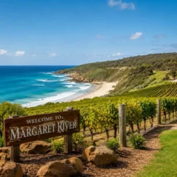 Scenic coastal view of Margaret River, Western Australia, featuring a wooden welcome sign in the foreground, lush green vineyards, sandy beaches, turquoise ocean waves, and rolling hills under a clear blue sky.